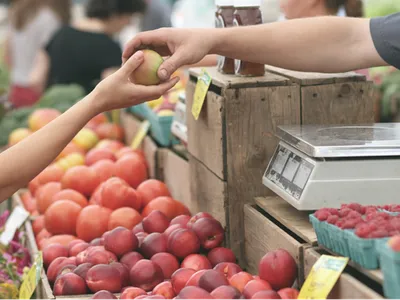 Hand passing a green apple at a produce stall with peaches, raspberries, and a scale.