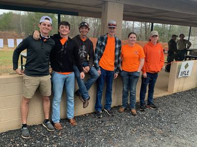 Six teens in orange shirts standing at an outdoor rifle range, sign "RIFLE"
