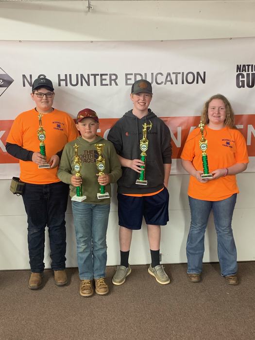 Four youths holding trophies, standing before banner reading "NO HUNTER EDUCATION"