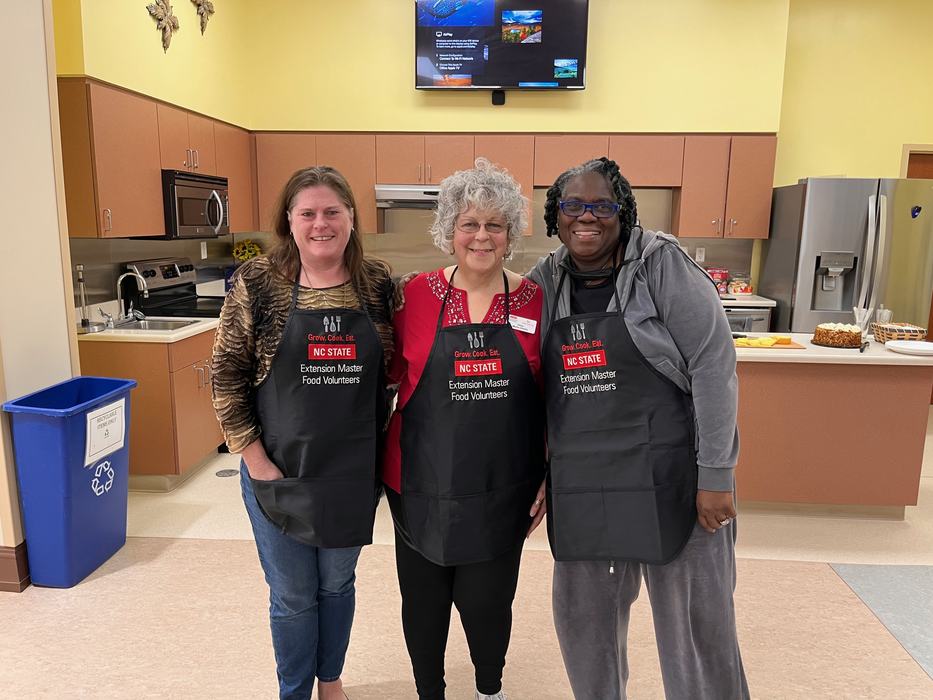 Three women in a kitchen wearing aprons reading "NC STATE Extension Master Food Volunteers."
