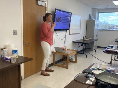 Woman standing by a door speaking to a classroom, TV monitor and laptop visible