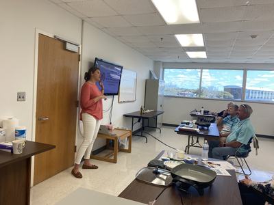 Woman standing by wall-mounted screen presenting to seated adults in a classroom