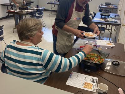 Man and woman cooking together in a classroom, man serving food onto a paper plate