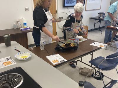 Two women in aprons serving cooked food from an electric skillet on a classroom table
