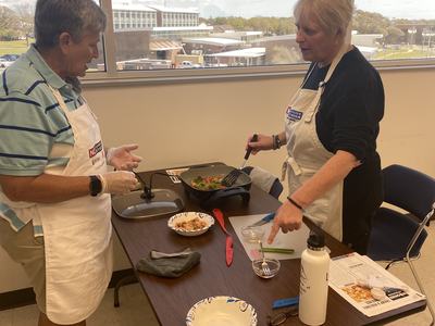 Two people in aprons cooking vegetables in an electric skillet on a table