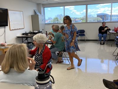 Community room with older adults seated at tables and one woman walking between them.