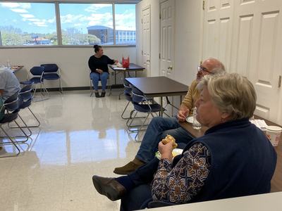 Three people seated in a breakroom; two eating at a table and one seated alone by a window.