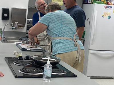 Three people in a kitchen preparing a fish on a cutting board; stovetop and sanitizer visible