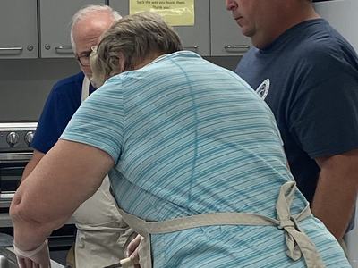Three people in a kitchen; a person in a striped shirt and apron filleting a fish on a counter
