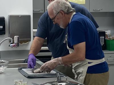 Two people in a kitchen wearing gloves and aprons filleting a fish on a cutting board.