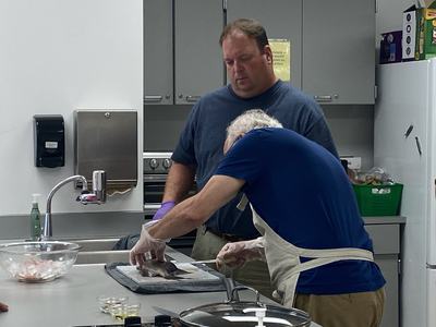 Two men in a kitchen, one filleting a fish on a countertop while the other watches