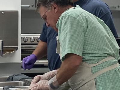 Two men in a kitchen wearing gloves and aprons preparing food on a countertop