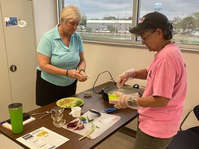 Two people preparing food at a table with cutting board, bowls, utensils, and ingredients.