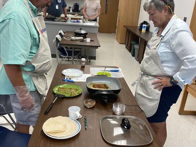 Two adults in aprons standing at a table with an electric griddle and tortillas.