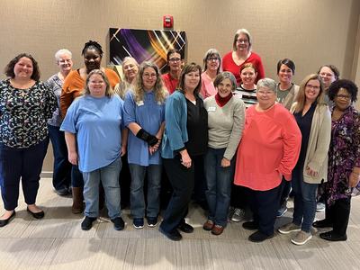 Group of 18 women standing in two rows indoors, posing for a group photo.