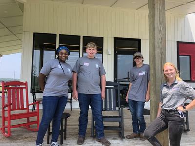 Four young people standing on a porch in gray shirts beside rocking chairs.