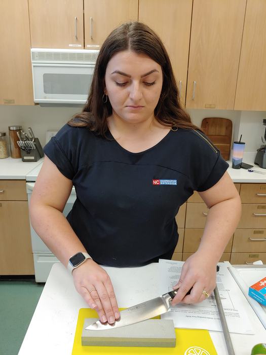 Person sharpening a chef's knife on a whetstone in a kitchen; shirt text "NC Cooperative Extension"