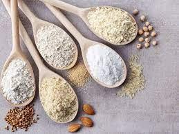 Five wooden spoons holding different flours and grains, with almonds and scattered seeds nearby