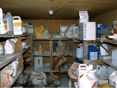 Storage room with wooden shelves holding chemical jugs, sacks, and a box labeled "UP"