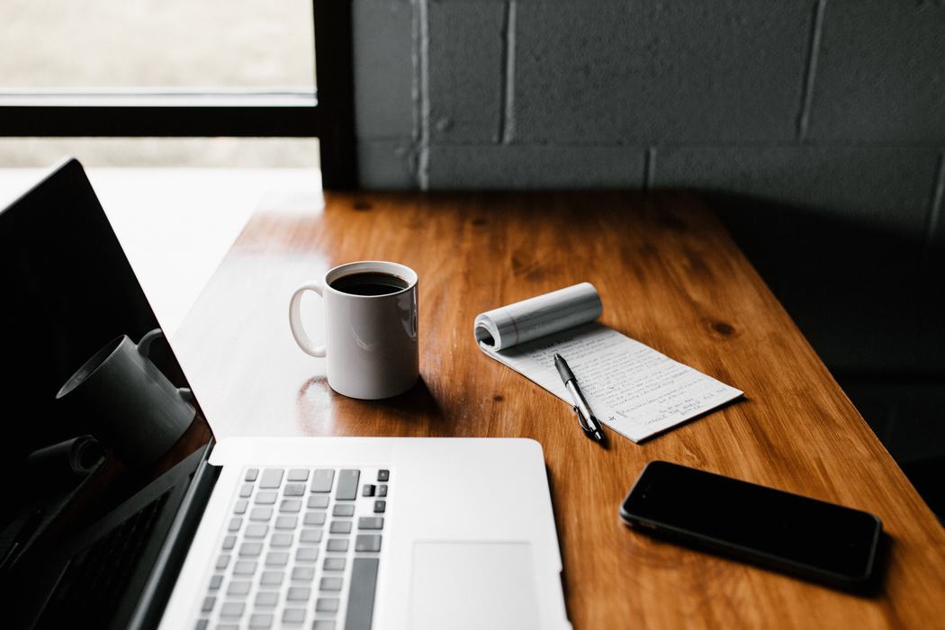 Laptop, coffee mug, notepad with pen, and smartphone on a wooden desk by a window