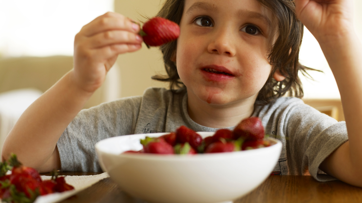Child holding a strawberry above a bowl of strawberries, red juice stains around mouth