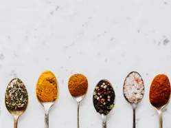 Six spoons holding different spices and salts on a marble surface.