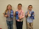 Three children holding blue ribbons, medals, and colorful gift bags against a wall