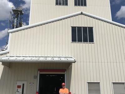 Man in orange shirt and red hard hat standing with arms crossed outside beige industrial building