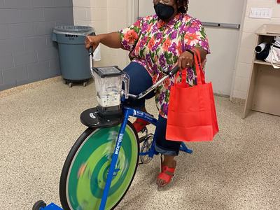 Person wearing floral shirt and mask pedaling a blender-mounted stationary bike under an "EXIT" sign