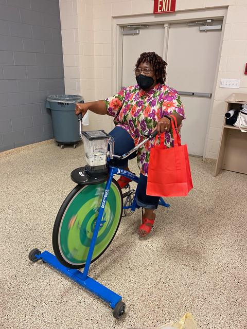 Person wearing floral shirt and mask pedaling a blender-mounted stationary bike under an "EXIT" sign