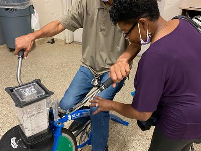 Man pedals blender bike while woman adjusts handlebars; blender jar labeled "Blendtec"