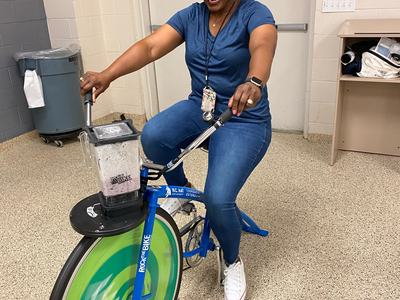 Woman pedaling stationary blender bike labeled "Rock the Bike" beneath an EXIT sign