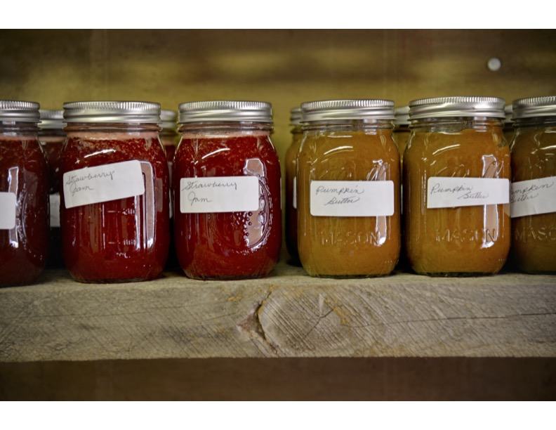 Row of mason jars labeled "Strawberry Jam" and "Pumpkin Butter" on wooden shelf