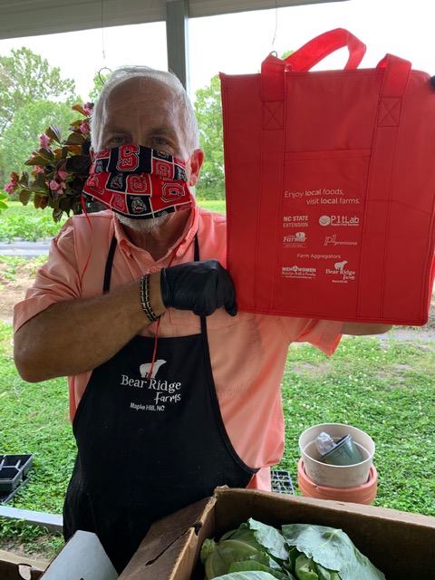 Person wearing mask and apron holds red bag reading "Enjoy local foods, visit local farms."