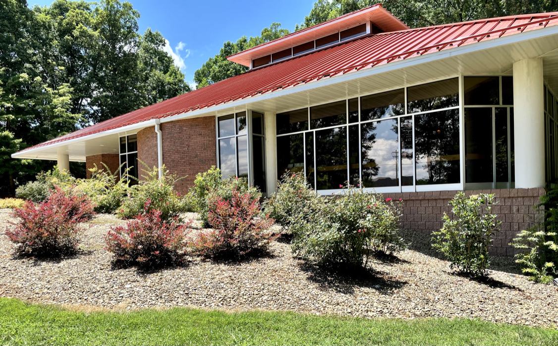 Single-story brick building with red metal roof and landscaped shrub beds