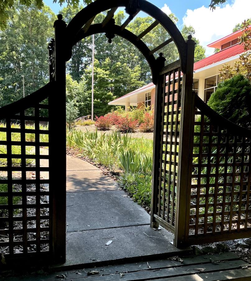 Open wooden garden gate overlooking walkway, flower beds, and a red-roofed building.