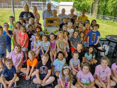 A group of first graders sit in front of a beehive.