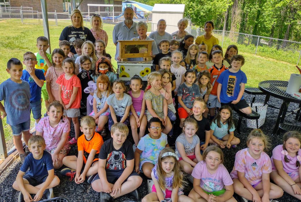 A group of first graders sit in front of a beehive.