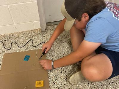 Person wearing cap and safety glasses soldering wires on cardboard on a speckled floor