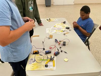 Three boys at a table assembling simple electrical circuits with batteries and wires