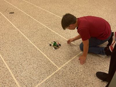 Child kneeling on speckled floor adjusting a small wheeled robot car on taped lanes
