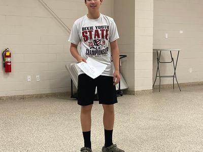 Teen standing in room holding papers, wearing T-shirt reading "DIXIE YOUTH STATE CHAMPIONSHIP"