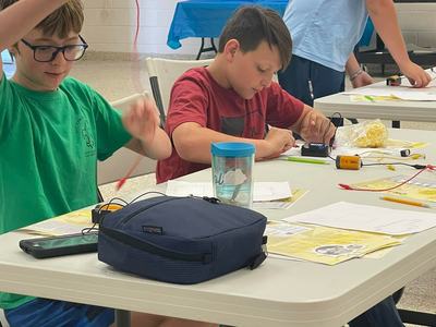Three boys at a table assembling electronics kits; one lifts a red wire, another connects components