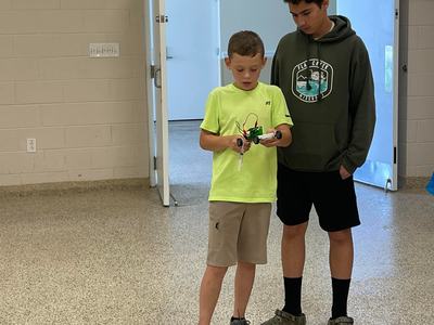 Two boys standing in a room; younger boy holds a small wheeled robot with exposed wires.