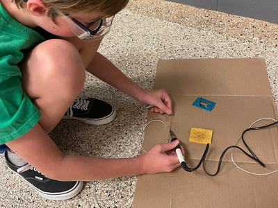 Child with safety goggles kneeling, using a soldering iron to join wires on cardboard.