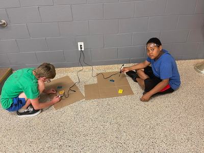 Two children sitting on floor soldering components on cardboard with plugged-in irons