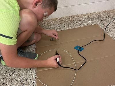 Child wearing safety goggles uses a soldering tool on a wire loop placed on cardboard