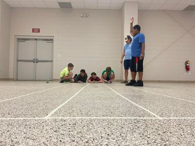 Six boys at marked lanes preparing toy cars; one boy standing with hand raised; 'EXIT' sign