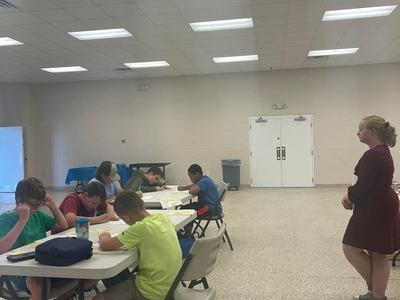 Several children seated at tables writing on papers while an adult stands nearby in a large room