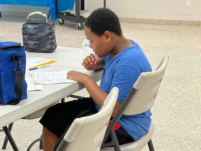 Child seated at folding table reading papers, pen and water bottle nearby.